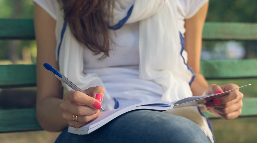 Young female student sitting on a bench in the park. She takes notes in a notebook. She is dressed in blue jeans and white T-shirt. Close-up. ** Note: Shallow depth of field
